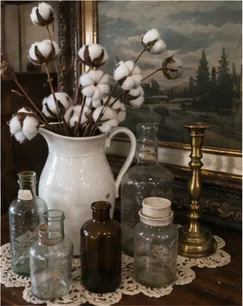Cotton-stem bouquet in a white pitcher with bottles and a brass candlestick
